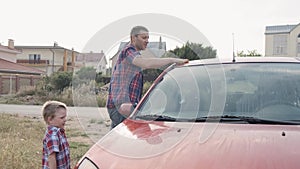 Young dad in red checkered shirt washes his car`s windshield window and the hood with his small son slow motion