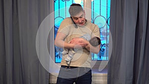 A young dad rocking a child in his arms against the background of the window. The man does not know what to do and