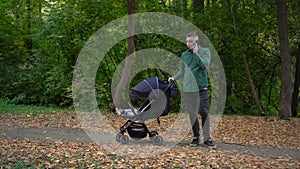 A young dad is talking on the phone and rocking his child in a stroller. A man speaks on the phone in the autumn forest.