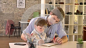 Young dad teaching his little son how to write, sitting at table in modern office