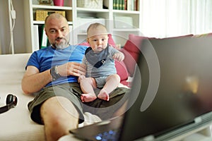 Young dad works remotely from home office with baby. Freelancer man holding his infant while using laptop. Workplace in living