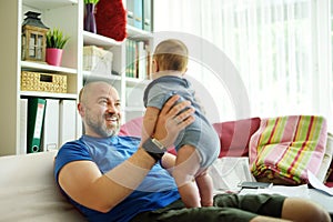 Young dad works remotely from home office with baby. Freelancer man holding his infant while using laptop. Workplace in living