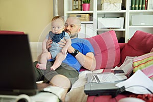 Young dad works remotely from home office with baby. Freelancer man holding his infant while using laptop. Workplace in living