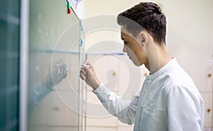 A young dark-haired Caucasian student stands near the blackboard and writes with chalk. A teenager is thinking at the