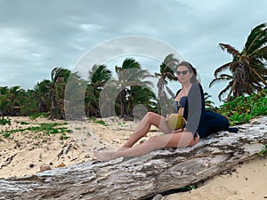 A young dark haired european girl with coconut in her hands is sitting on a fallen tree on the white sand Caribbean beach in