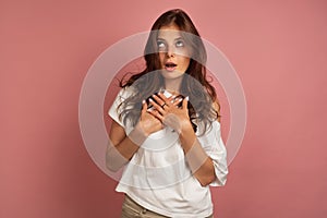 Young dark-haired girl in a white t-shirt puts her hands to her chest as if she expresses amazement, pink background