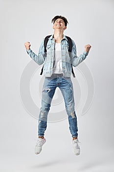 Young dark-haired guy with black backpack on his shoulder dressed in a white t-shirt, jeans and a denim jacket happily jumps on