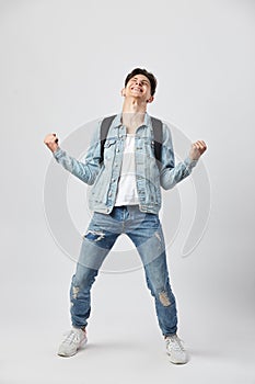 Young dark-haired guy with black backpack on his shoulder dressed in a white t-shirt, jeans and a denim jacket looks