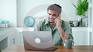 Young dark-haired male entrepreneur using laptop in the home kitchen interior, talking on phone with business partners