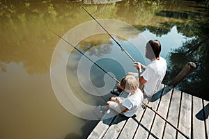 A young dark-haired man and a blond boy dressed in the white t-shirts are sitting with fishing rods on the wooden pier