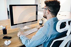 Young dark-haired man is working with a computer at his desktop in office. He wears blue shirt and looks busy. View from