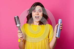Young dark haired woman wears yellow shirt holds hair brush and hair spray. Attractive lady looks surprised. Female with rose