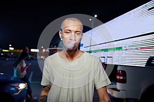 A young dark-skinned well-dressed tattooed man looking into a camera while breathing out smoke having friends and a led