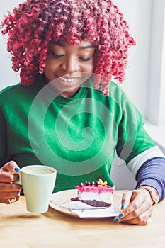 Young dark-skinned woman dressed in casual clothes holding cup of hot drink, enjoying coffee and cheesecake