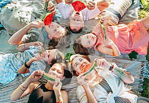 Young daughters with parents family lying on picnic blanket during weekend sunny day, smiling, laughing and eating red juicy