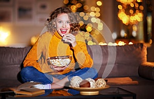 Young  delighted cheerful woman with popcorn laughs and watches  christmas movie on  TV   at home in evening  alone