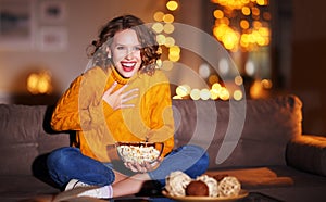 Young  delighted cheerful woman with popcorn laughs and watches  christmas movie on  TV   at home in evening  alone photo