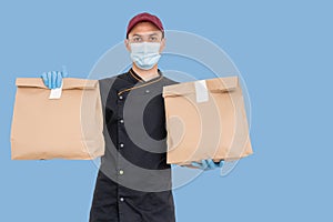 Young delivery man in medical mask holding and carrying a cardbox isolated on blue background. Buy food online in