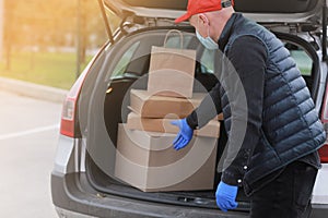 Young delivery man in protective mask, red cap and gloves near the car with boxes and packages, outdoors. Service coronavirus.