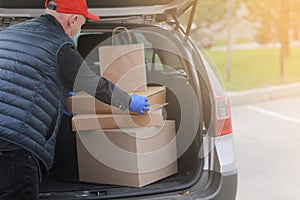 Young delivery man in protective mask, red cap and gloves near the car with boxes and packages, outdoors. Service coronavirus.