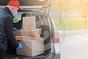 Young delivery man in protective mask, red cap and gloves near the car with boxes and packages, outdoors. Service coronavirus.