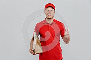 Young delivery man in uniform showing thumb up while holding paper bag