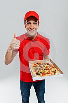 Young delivery man in uniform showing thumb up while showing pizza