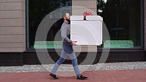 Young delivery man walking along city street, carrying in hands surprising festive cardboard box.
