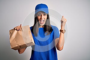 Young delivery woman with blue eyes wearing cap holding paper bag with food screaming proud and celebrating victory and success