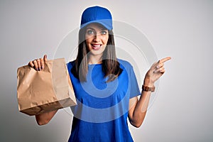 Young delivery woman with blue eyes wearing cap holding paper bag with food very happy pointing with hand and finger to the side