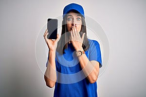 Young delivery woman with blue eyes wearing cap holding smartphone cover mouth with hand shocked with shame for mistake,