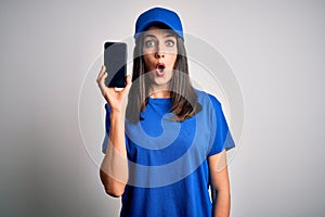 Young delivery woman with blue eyes wearing cap holding smartphone scared in shock with a surprise face, afraid and excited with
