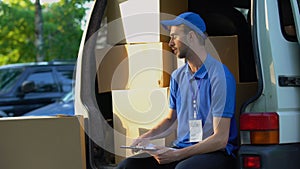 Young deliveryman writing parcel distribution report and smiling, sitting in van