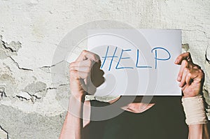 Young depressed homeless man with bandage on his hand from suicide attempt holding help sign written on paper while he leaning his