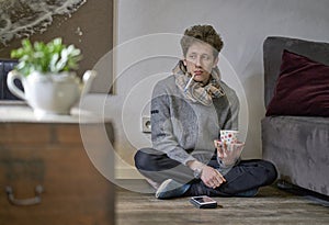 Young depressed looking man with a cold sitting on the floor in his apartment with a thermometer in his mouth and a cup of tea in