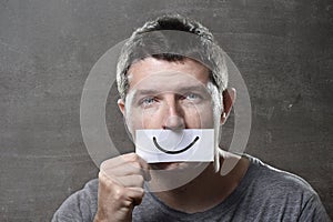 Young depressed man lost in sadness and sorrow holding paper with smiley on his mouth in depression concept