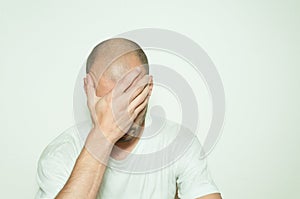 Young depressed man suffering from anxiety and feeling miserable cover his face with his hands and leaning on the white wall.