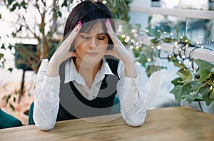 Young depressed woman sits in a cafe behind a wooden steel, holds her head in her hands, has a headache, low or high