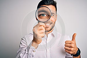 Young detective man looking through magnifying glass over isolated background happy with big smile doing ok sign, thumb up with