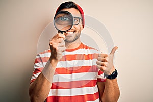 Young detective man looking through magnifying glass over isolated background happy with big smile doing ok sign, thumb up with