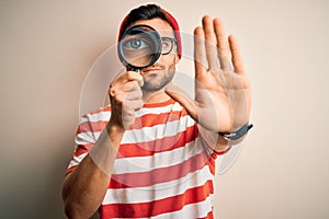 Young detective man looking through magnifying glass over isolated background with open hand doing stop sign with serious and