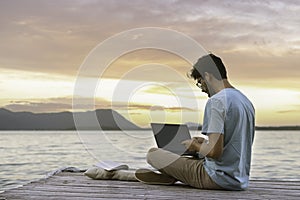 Young digital nomad man sitting on wooden pier at sea working on internet remotely at sunset - Traveling with a computer - Online