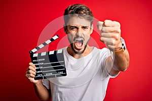 Young director man with beard making movie using clapboard over isolated red background annoyed and frustrated shouting with