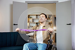 Young disabled man in wheelchair doing exercises with rubber band at home. Caucasian handicapped guy working out in living room