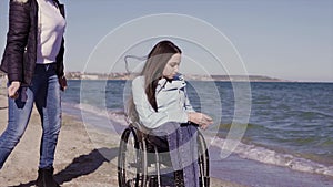 Young disabled woman in wheelchair with her mother having fun with stone skipping on the sand beach near the sea