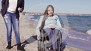 Young disabled woman in wheelchair with her mother having fun with stone skipping on the sand beach near the sea