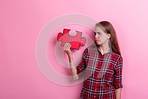 Young disgruntled girl, holds a big puzzle and looks at it. On a pink background.