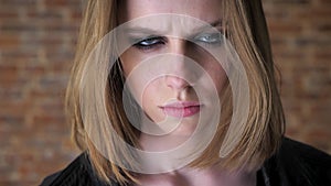 Young disgusted girl with smoky eyes is watching at camera, calming down, brick background, blurred background