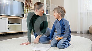 Young displeases mother shouting and talking strictly to her son doing homework on carpet at living room. Concept of