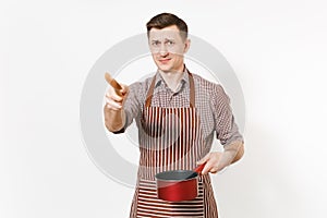 Young dissatisfied man chef or waiter in striped brown apron, shirt holding red empty stewpan, wooden spoon isolated on